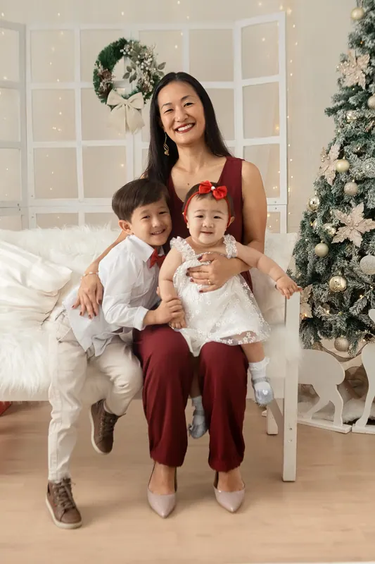Mãe sorridente sentada com os dois filhos ao lado de árvore natalina iluminada