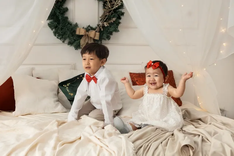 Irmãos sorriem em cama decorada com guirlanda e luzes no ensaio de Natal